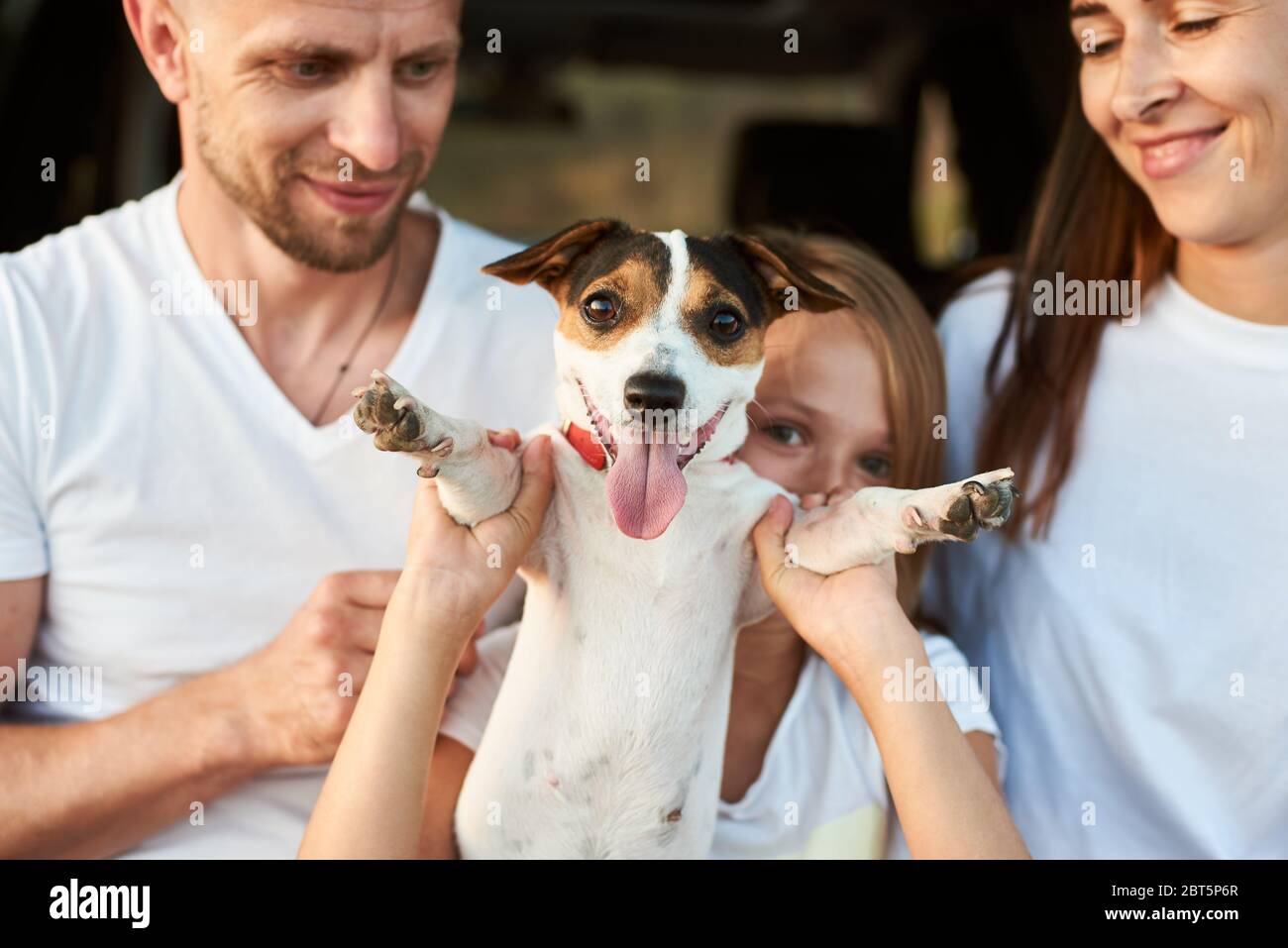 Close-up shot focused on jack russell terrier held by paws a girl, father and mothers are smiling on the blured background, white family look. Dog looking to the camera Stock Photo