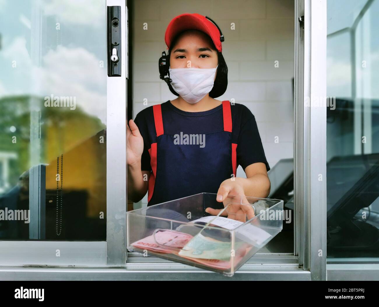 fast food cashier in drive thru service waring hygiene face mask to protect coronavirus pandemic or covid-19 virus outbreak is giving money change to Stock Photo