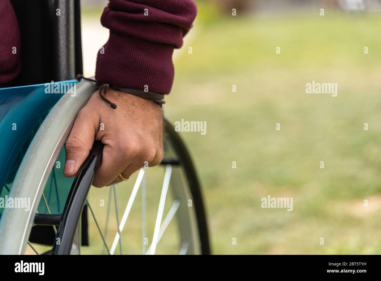 Hand of a paralyzed adult man in a wheelchair wearing a dark red sweatshirt in a park Stock Photo