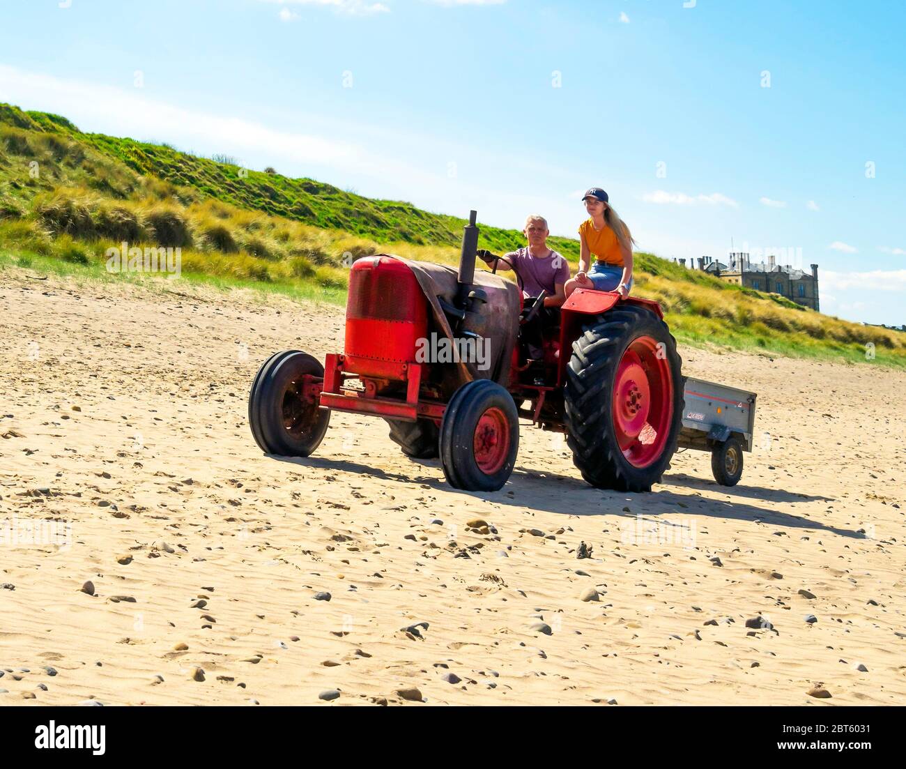 A white Caucasian man driving a red tractor along a sandy beach in North Yorkshire with a young woman with a long pony tail riding as a passenger Stock Photo