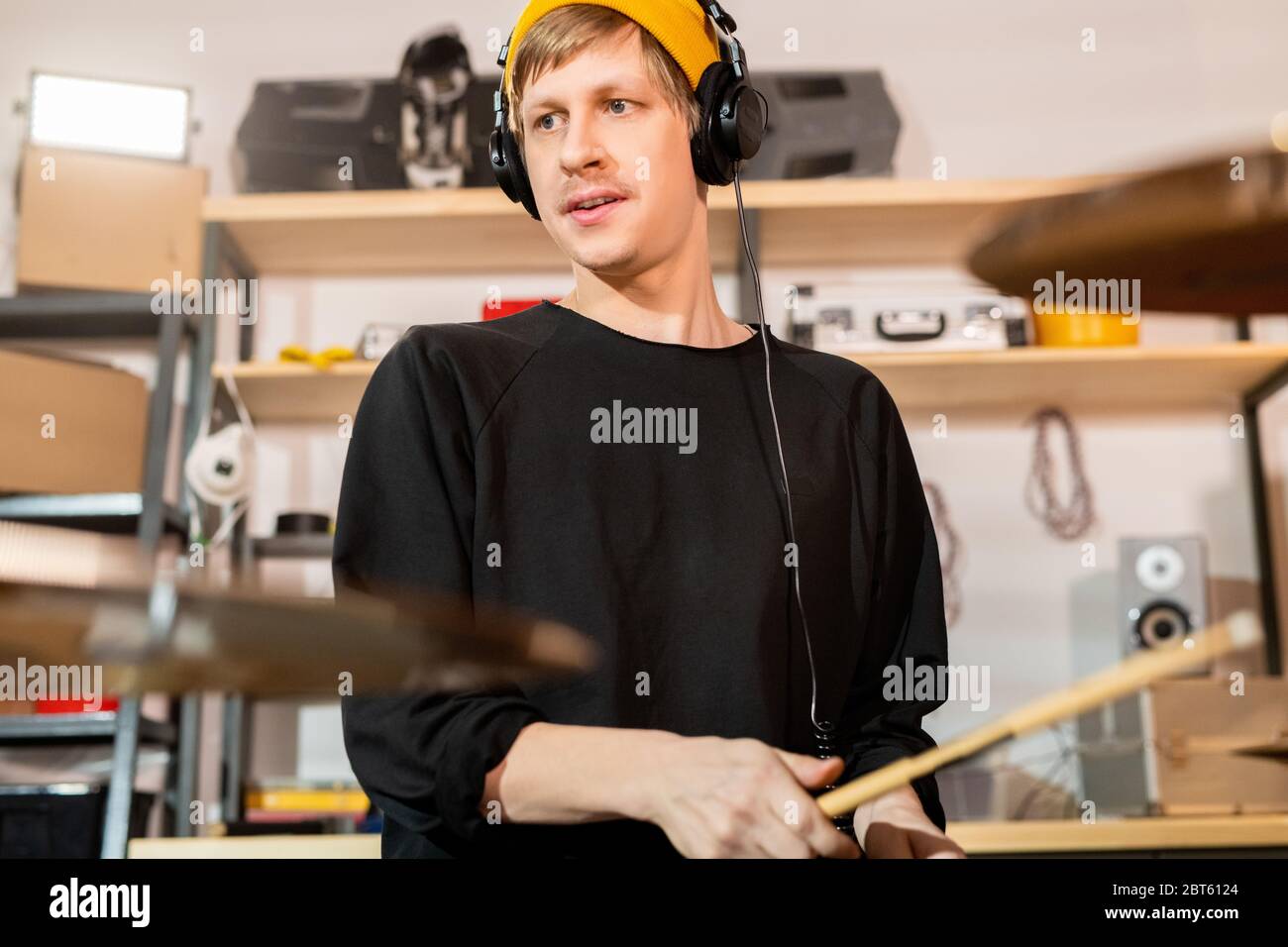 Contemporary young blond male drummer in headphones, yellow beanie and black sweatshirt hitting cymbals and drum in garage Stock Photo