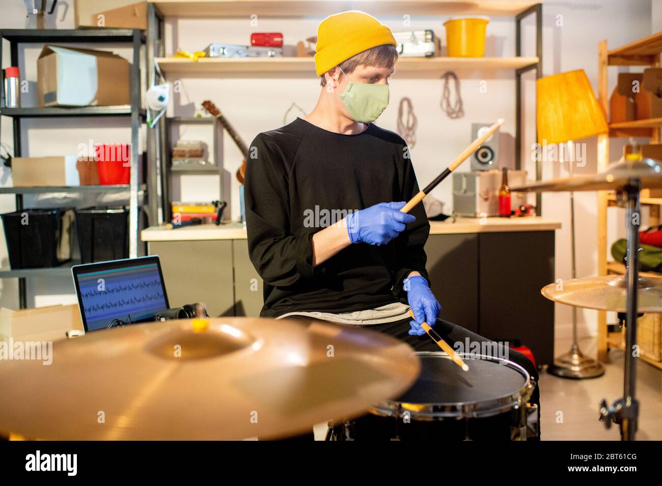 Young man in casualwear and protective mask and gloves hitting cymbals and drums while sitting in front of drumset and recording music Stock Photo