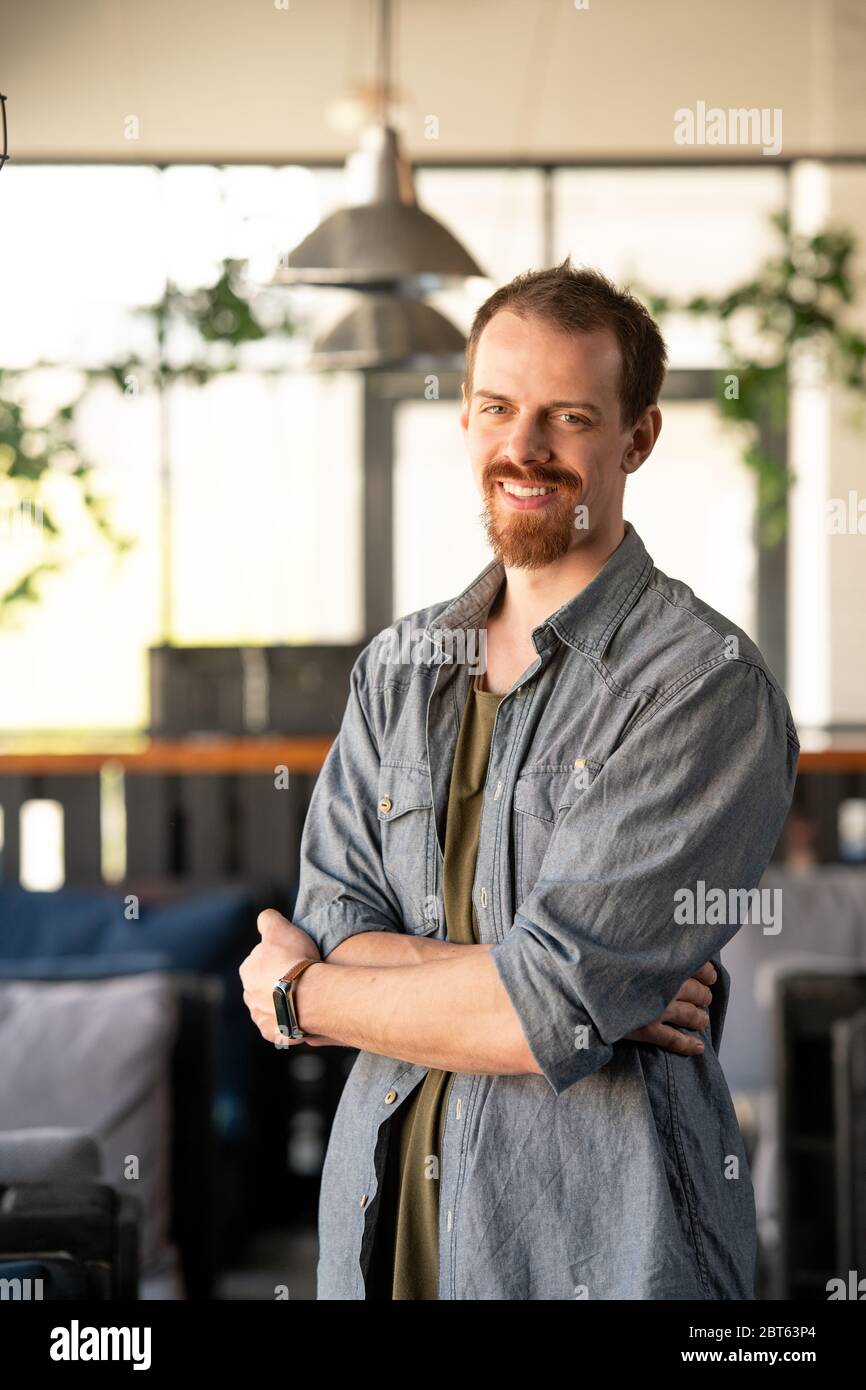 Successful young businessman with toothy smile crossing arms by chest while standing in front of camera in modern cafe environment Stock Photo