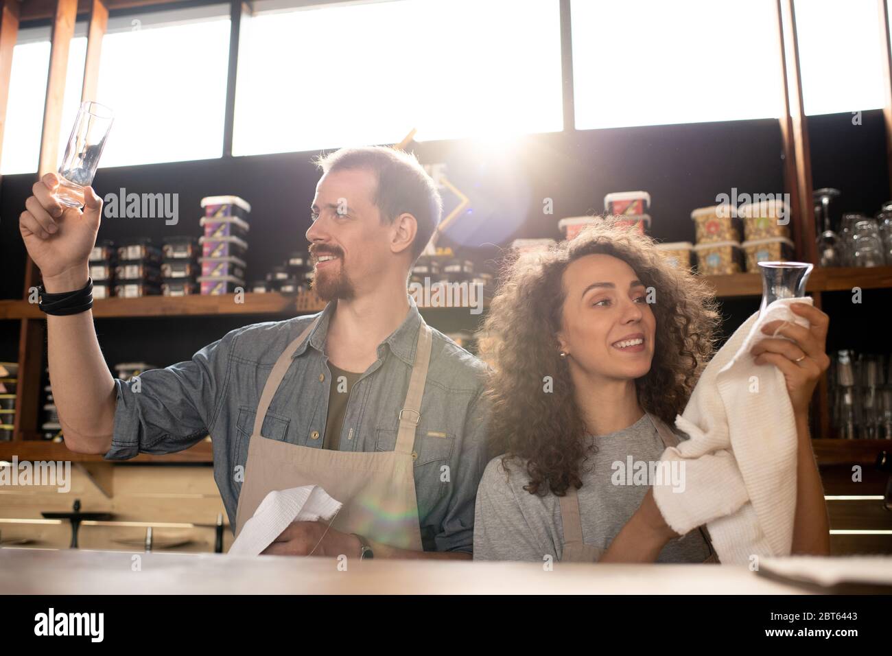 Happy young owners of cafe or restaurant standing by counter and cleaning glasses with towels at the end of working day Stock Photo