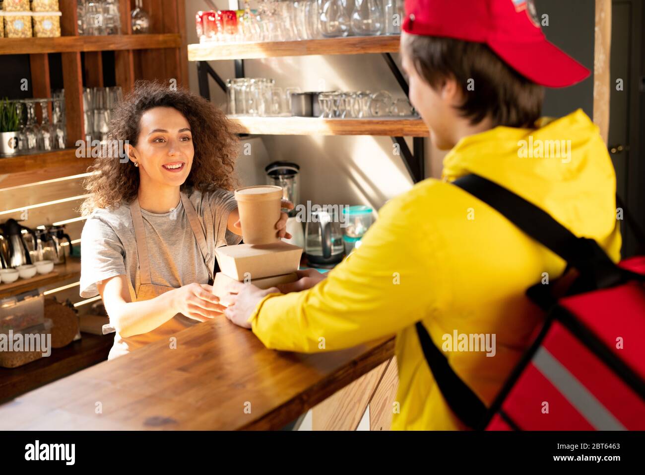 Happy young waitress of restaurant or cafe putting glass of coffee on top of carton containers with snack while talking to courier Stock Photo