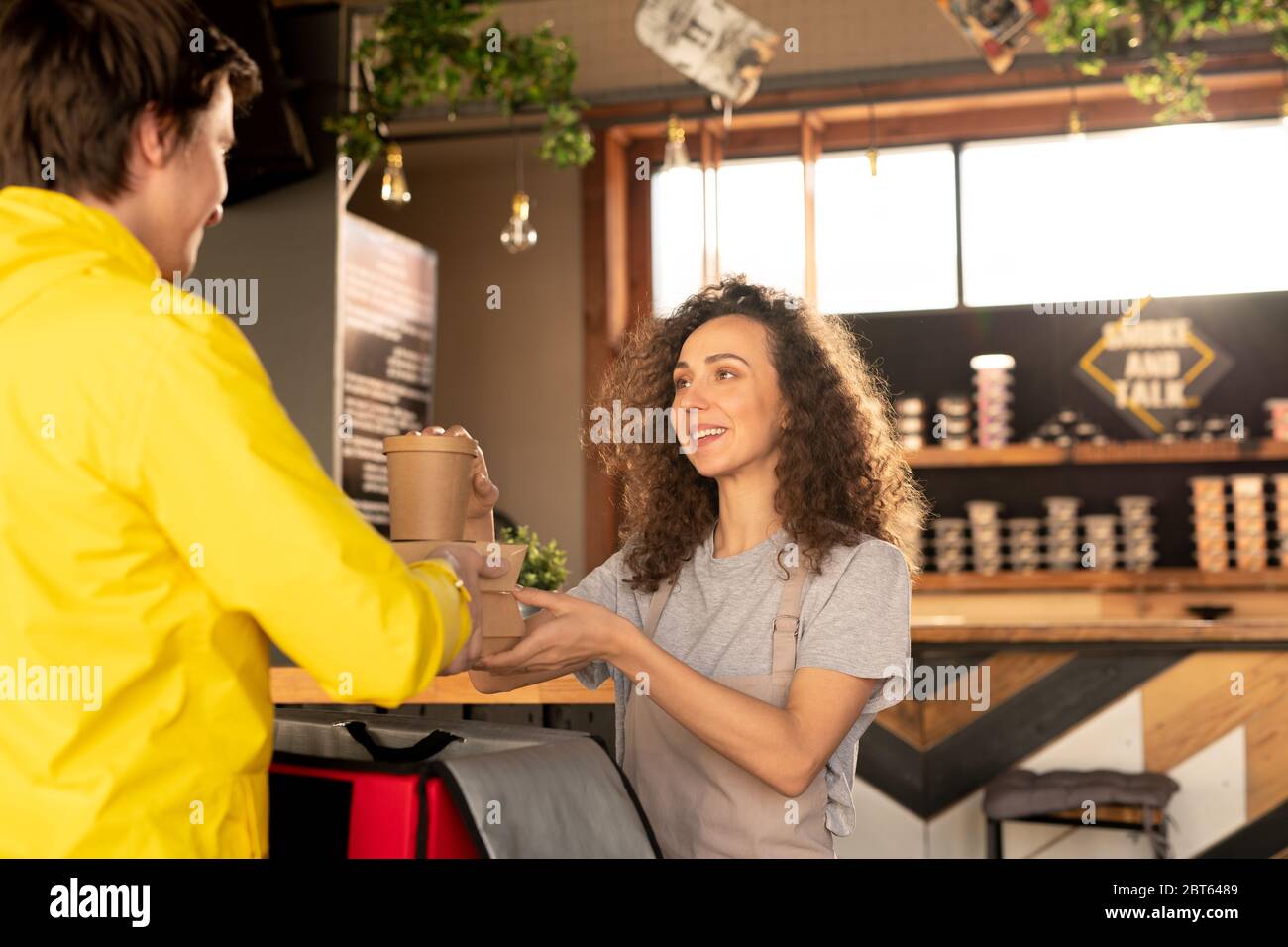 Young friendly waitress passing courier containers with ordered food and drinks while looking at him and helping with packing bag Stock Photo