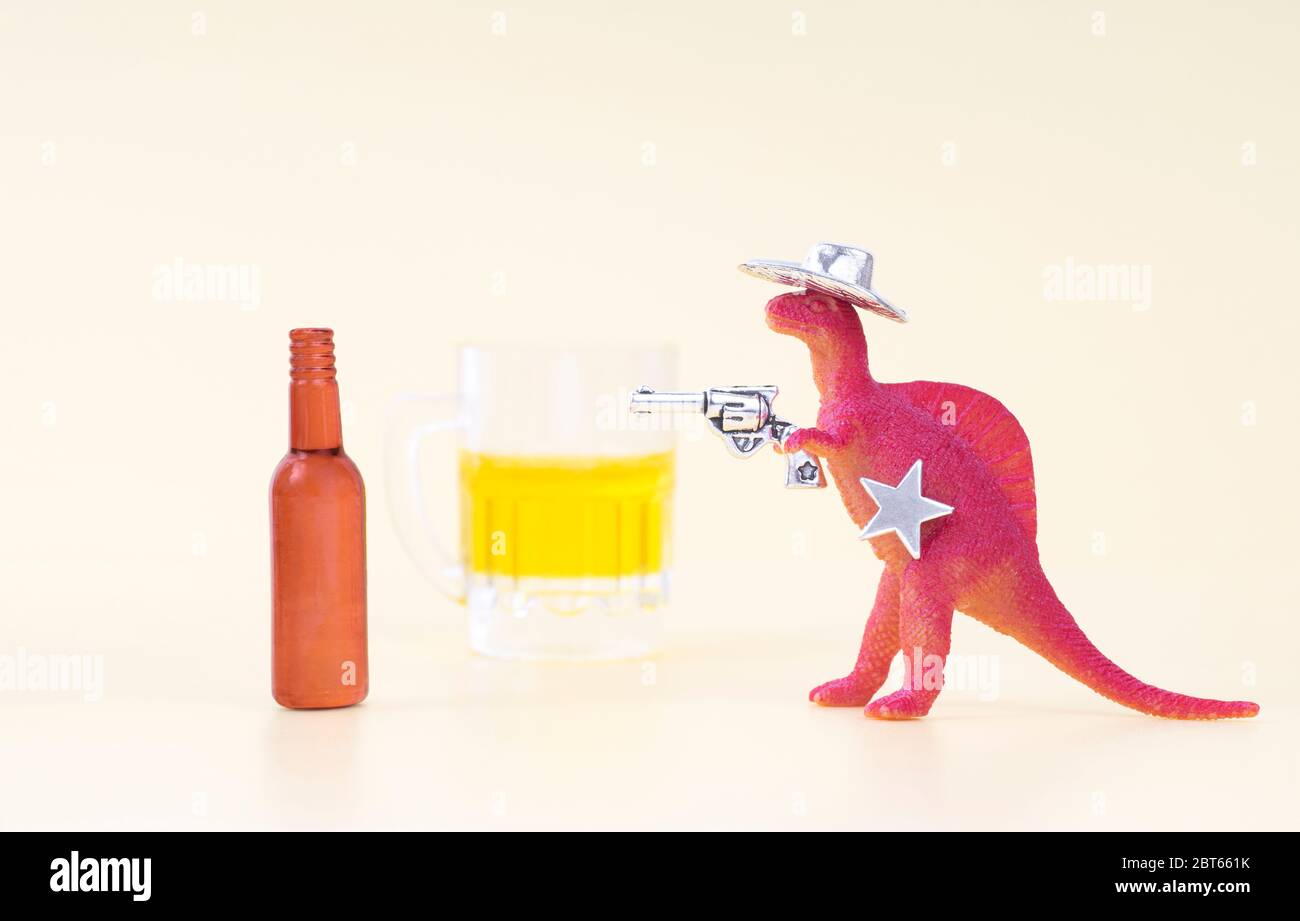 Toy dinosaur wearing a cowboy hat, holding a revolver in his arm and having a sheriff star shoots an empty beer bottle on a neutral background. Fight Stock Photo