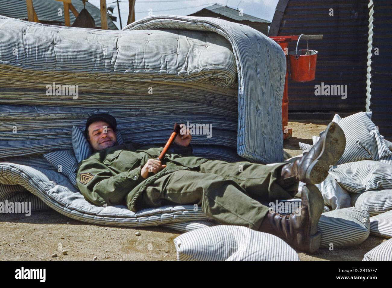 Enjoying off-duty barrack life for the US Army's 40th Infantry, Division Replacement Company in the Japan 1955, shortly after the Korean War was over. This smiling sergeant lies relaxed on a pile of mattresses and holds a baton (stick or truncheon). The 40th Infantry Division ('Sunshine Division') is part of the US Army.  It saw active service in the Korean War (1950–53), participating in the battles of Sandbag Castle and Heartbreak Ridge. The division remained in Korea until May 1954. In 1953 their HQ moved to Tokyo, then to Yokohama and finally in October to Camp Zama, southwest of Tokyo. Stock Photo