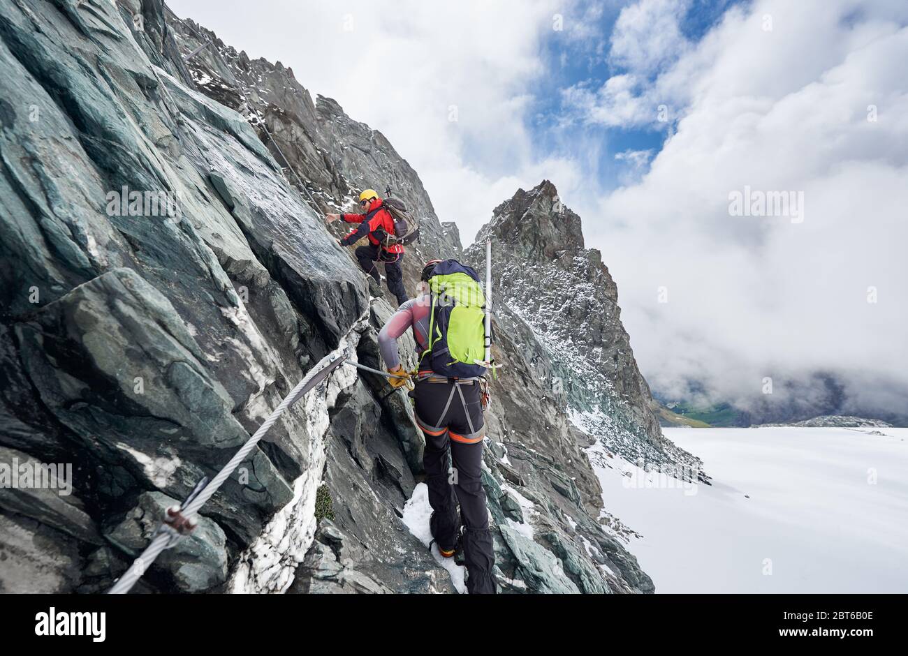 Back view of rock climbers with backpacks using fixed rope while ascending high rocky mountain. Male mountaineers climbing natural rock formation and trying to reach mountaintop. Stock Photo