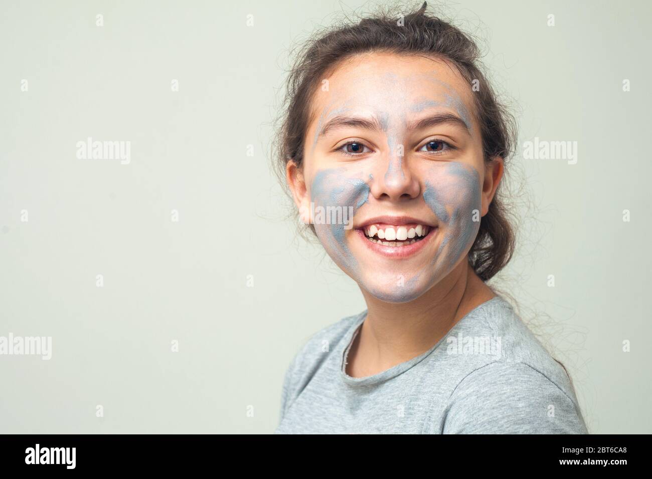 Cute and happy girl smears face with cosmetic clay or mud and smiles. Cosmetic mask, face scrub. The concept of health and beauty. Stock Photo