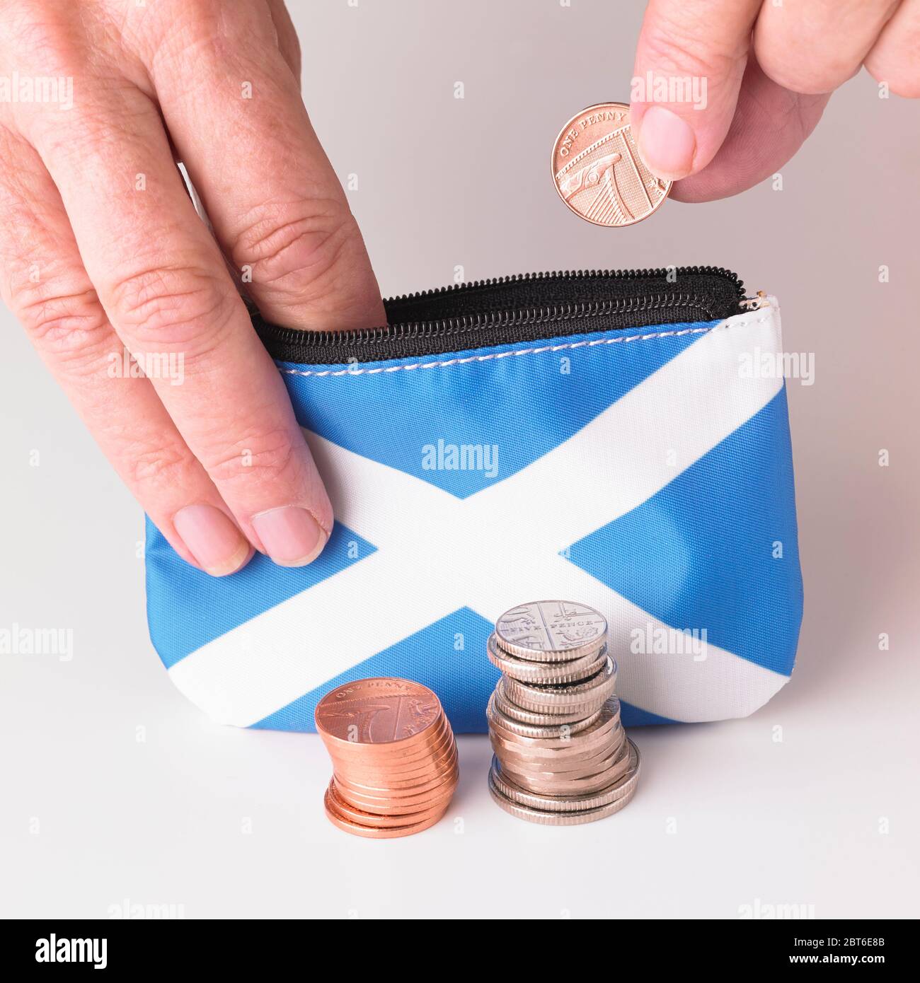 Hand placing British one penny coin into Scottish Saltire flag purse with two piles of mixed sterling coins in foreground Stock Photo