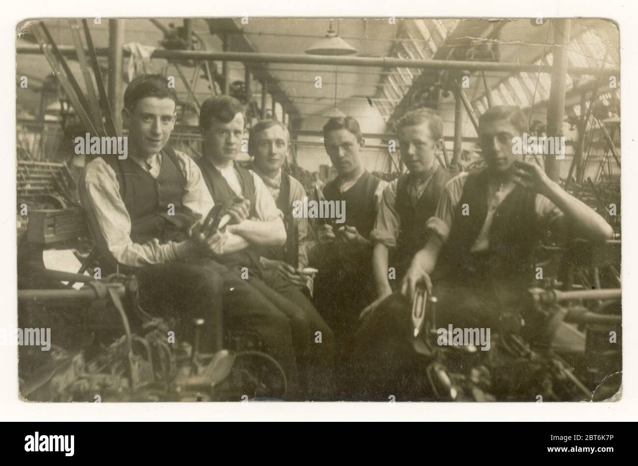 Original early 1900's postcard of group of young male cotton mill workers sitting next to loom machines, showing interior interiors of mill one of the lads is holding a 'flying shuttle' suggesting they are weavers, Blackburn, Lancashire, North West England, U.K. circa 1905 Stock Photo