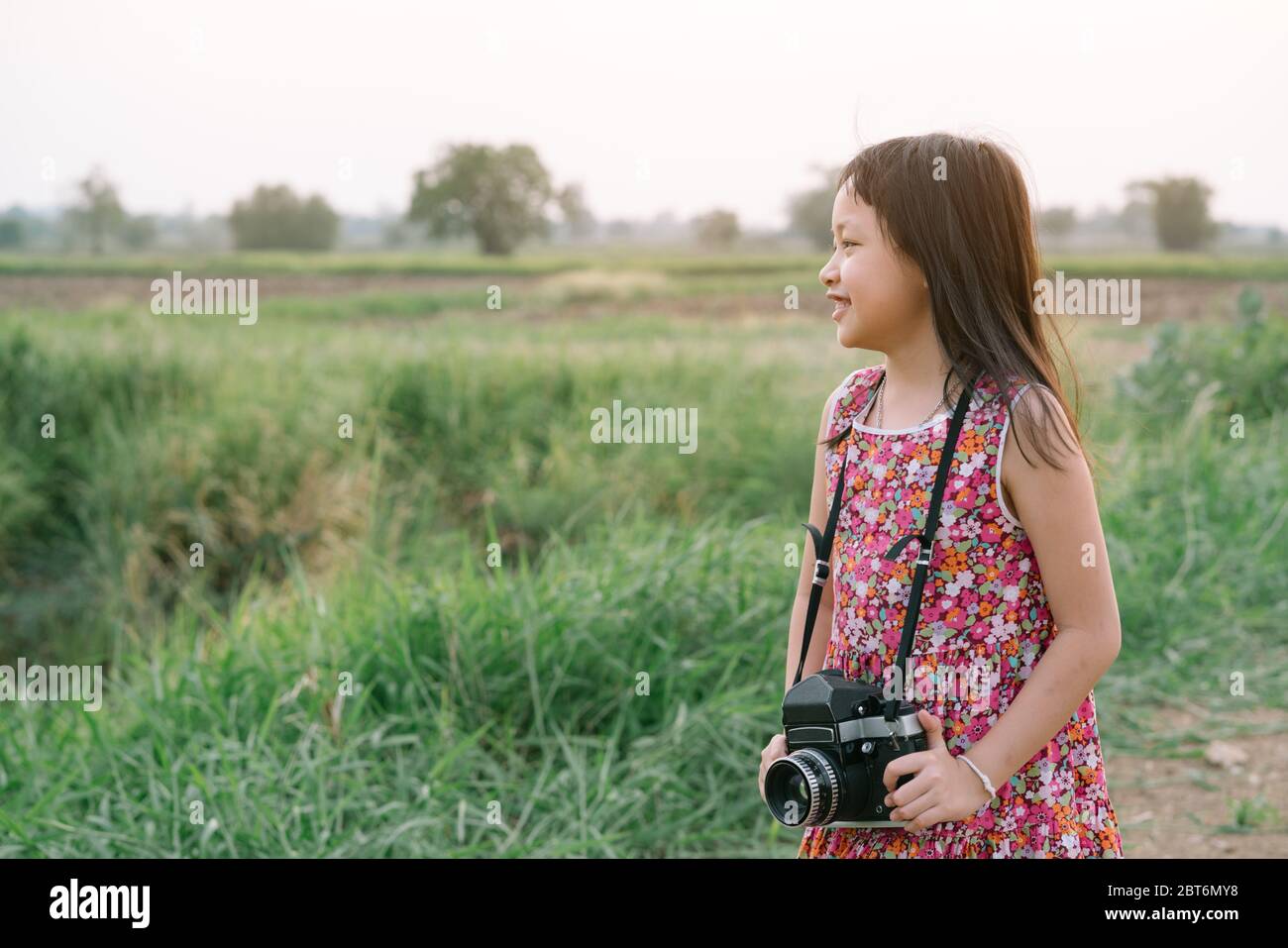 Little child girl holding medium format film camera and taking photo of sunset landscape with green field background Stock Photo