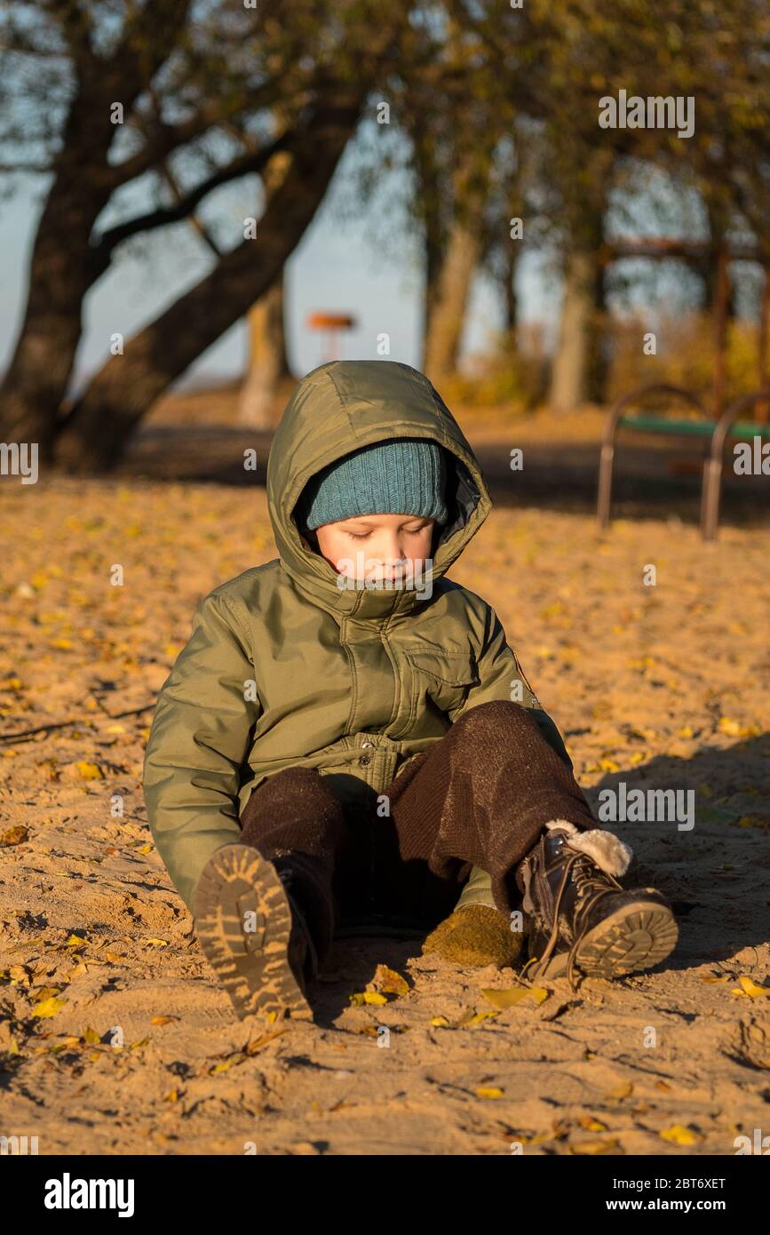 a boy in a green jacket, a blue knitted hat, brown trousers and boots stands on the yellow sand and looks down, stained his pants in the sand, Stock Photo