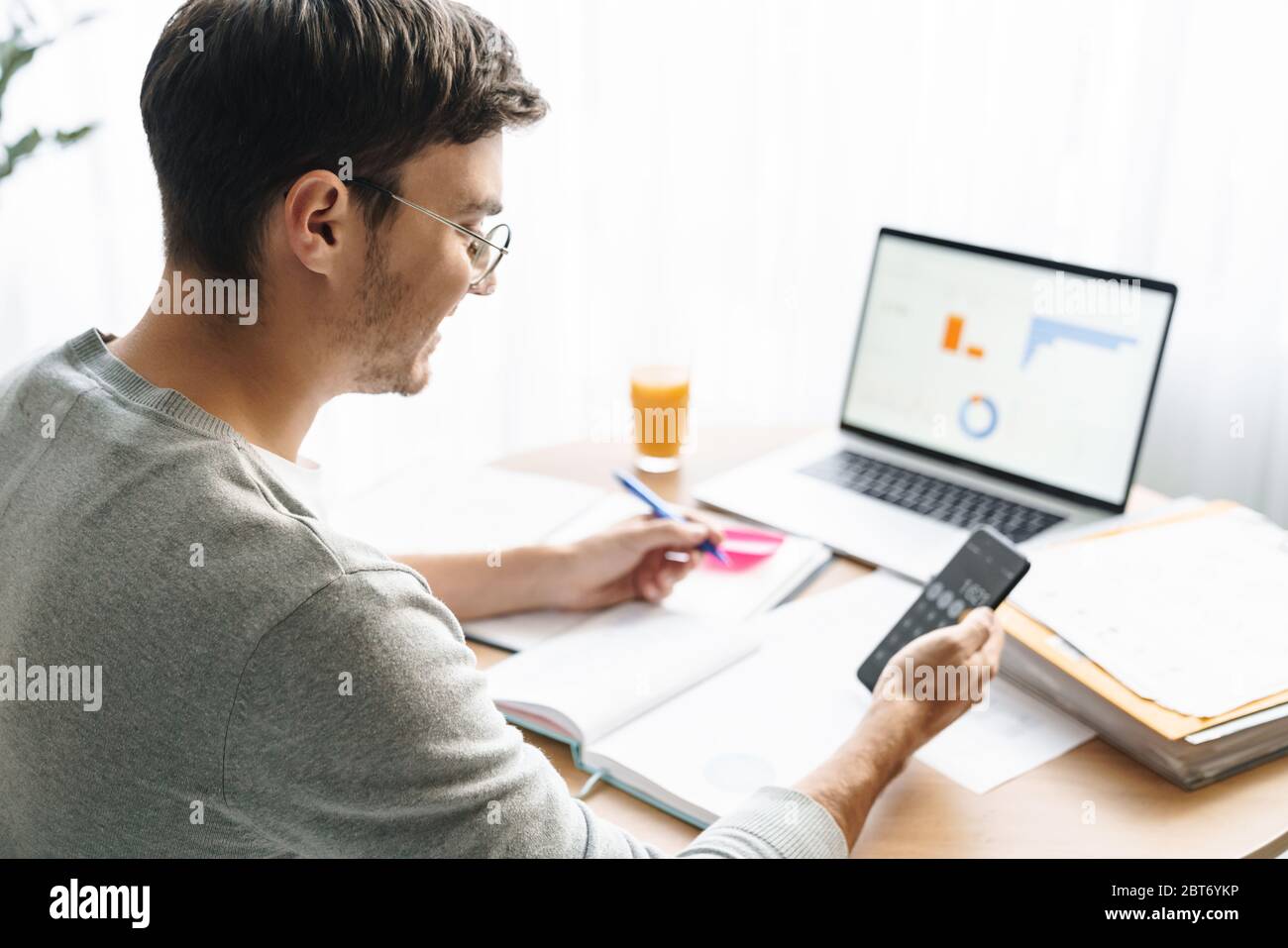 Image of happy young man in eyeglasses doing calculations while working on laptop at home Stock Photo