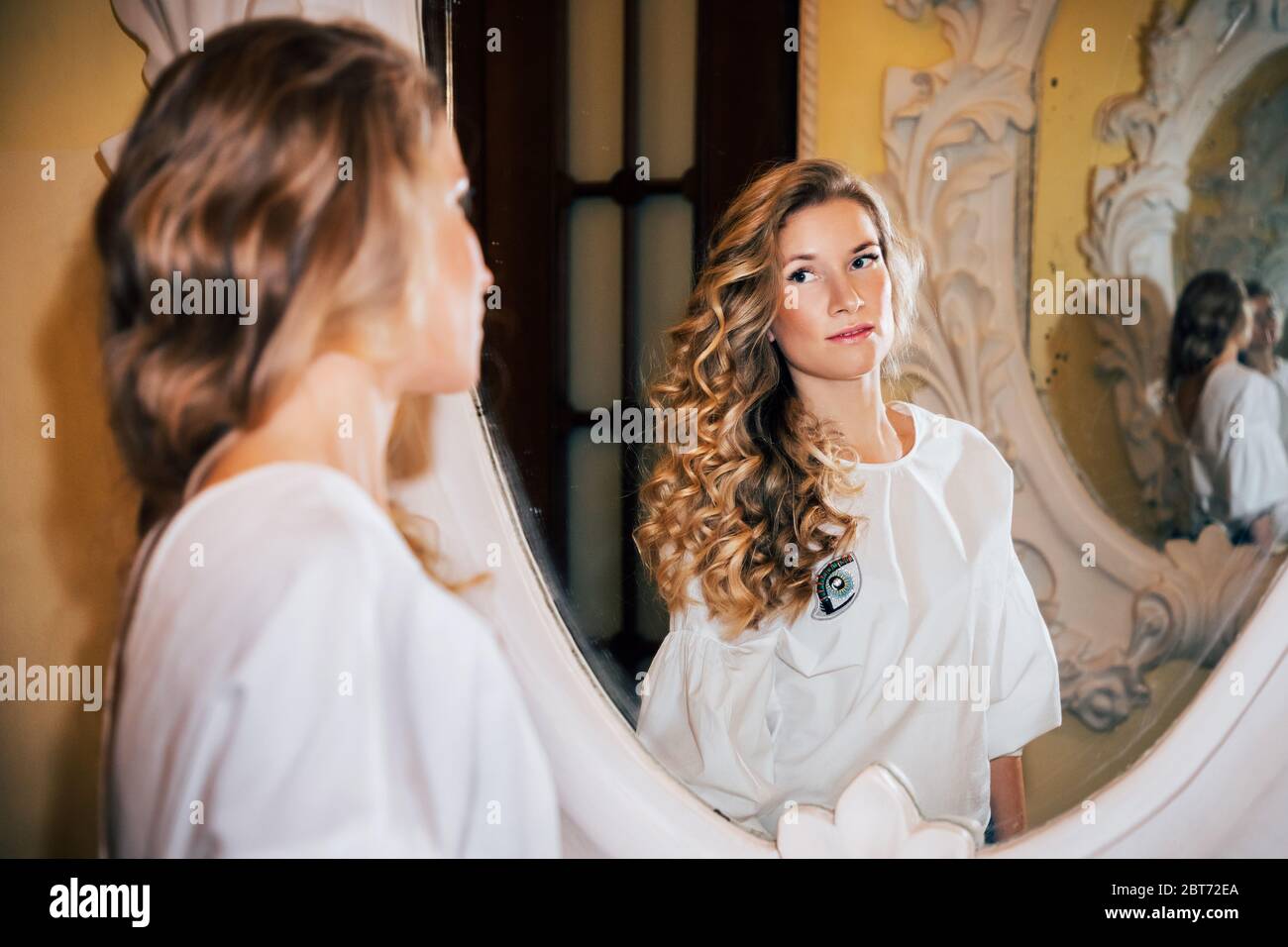Beautiful girl with long curly blonde hair, wearing a light top, is looking at herself in a richly ornamented mirror. Stock Photo