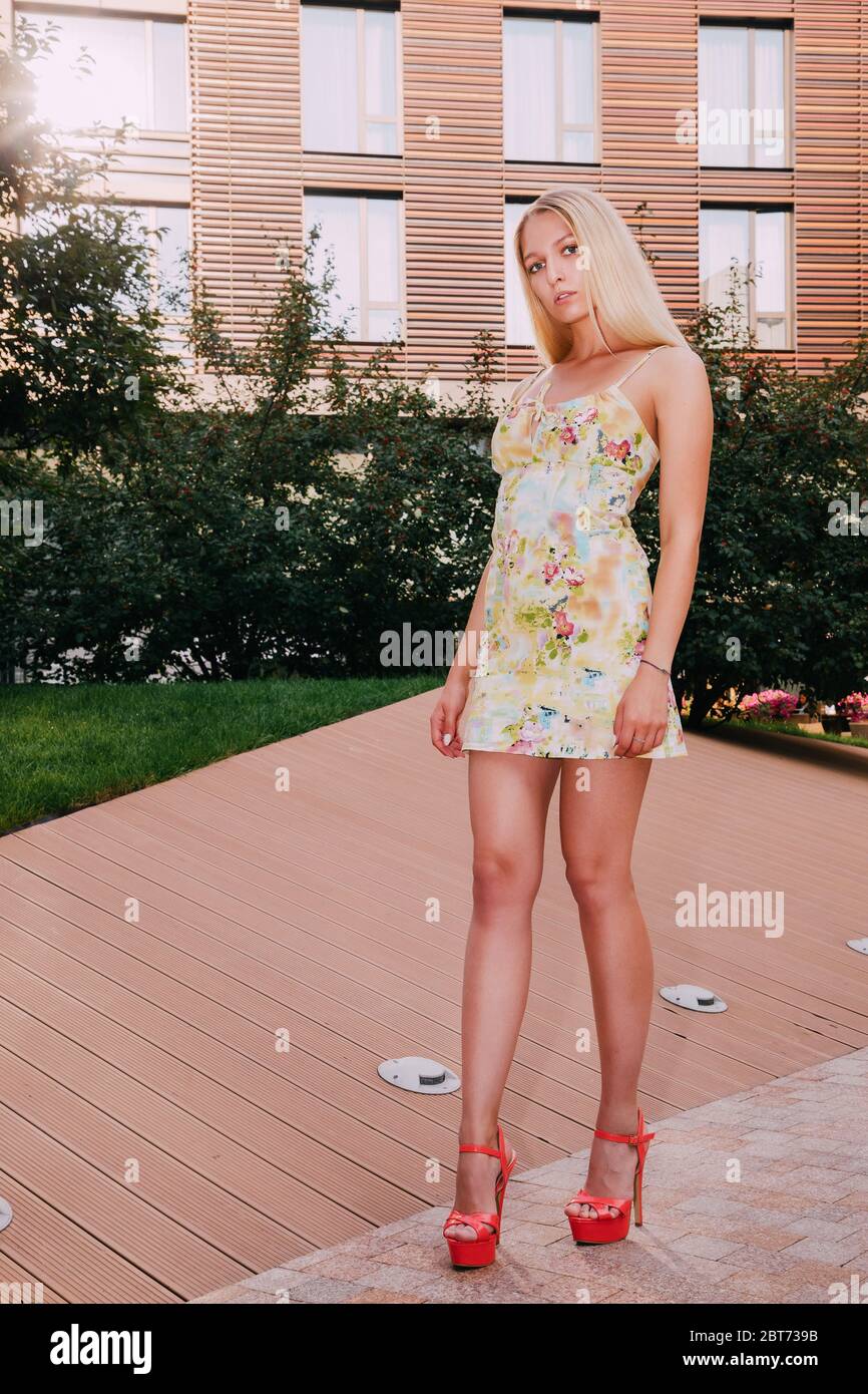 Pretty blonde girl is standing in front of a modern building. She is wearing a light short summer dress and red high heels. Stock Photo
