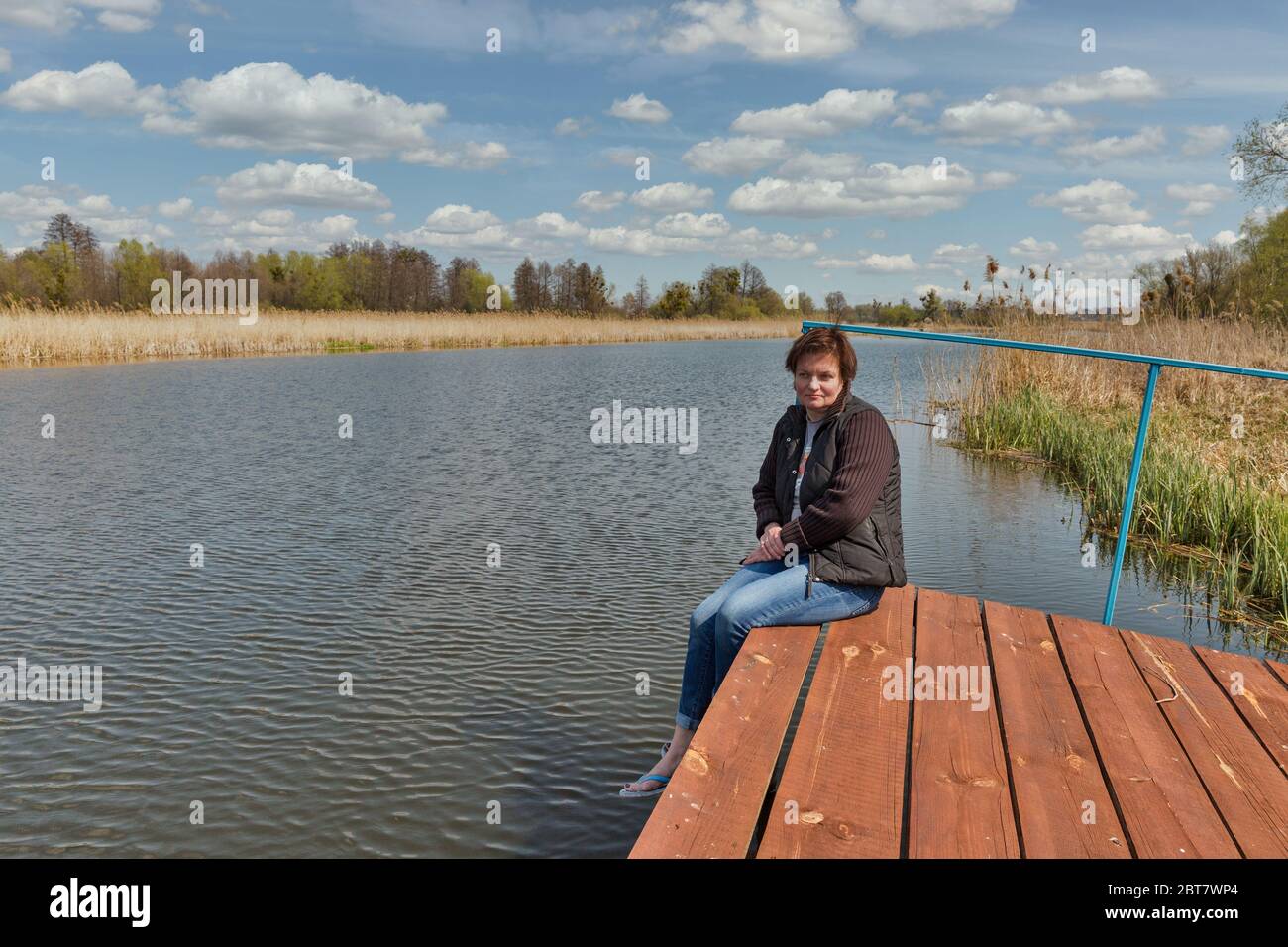 Caucasian middle aged woman facing at the river Ros while sitting on a wooden pavement and hugging her legs, Ukraine. Stock Photo