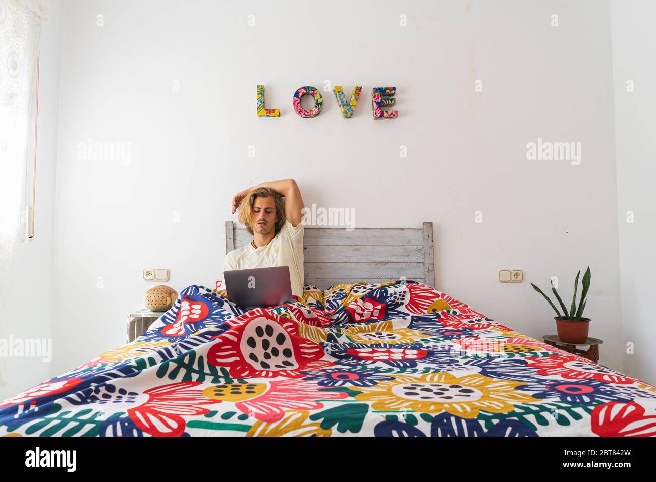 Young man with medium long blond hair, with a beard and mustache working with the computer from home during quarantine, from bed. Concept adapt to the Stock Photo