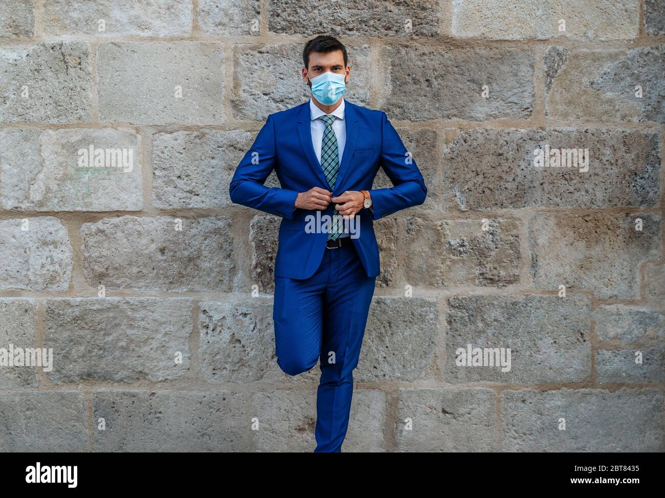 Young and stylish business man with face mask stands on old stone wall Stock Photo
