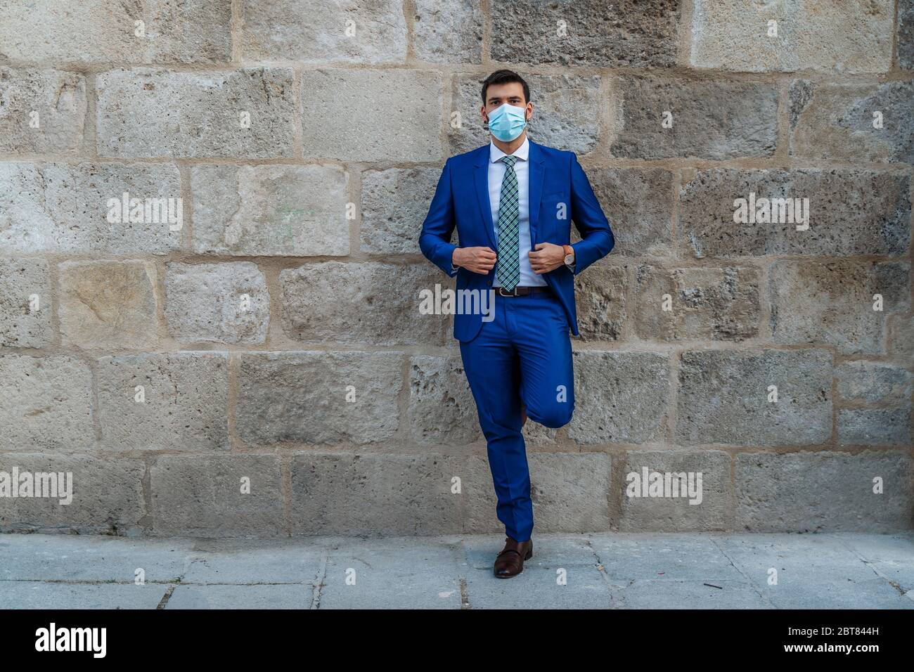 Young and stylish business man with face mask stands on old stone wall Stock Photo