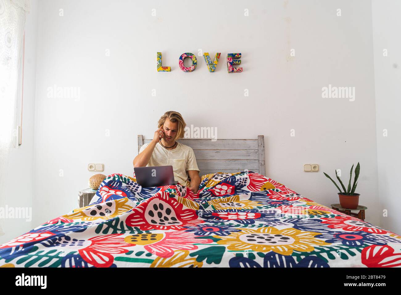 Young man with medium long blonde hair, with a beard and mustache working on the computer from home during quarantine, from bed. Calling by phone. Con Stock Photo