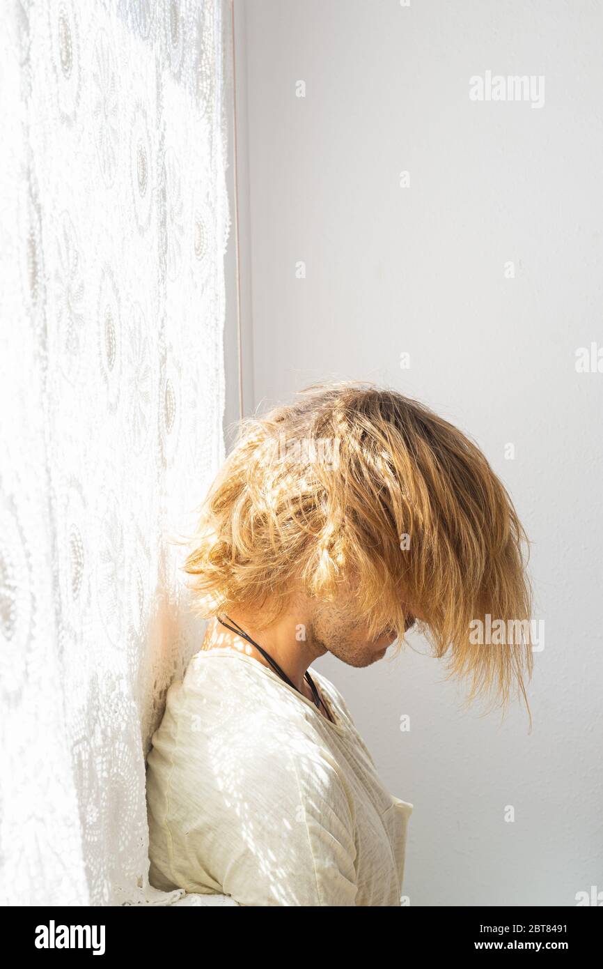 Young man with medium long blonde hair frustrated at not being able to leave home. Looking down on a white background. Loneliness and silence concept. Stock Photo