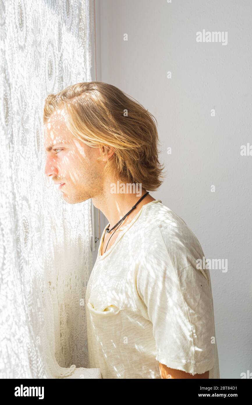 Young man with medium long blonde hair frustrated at not being able to leave home. Loneliness and silence concept. Sun rays coming into the house thro Stock Photo