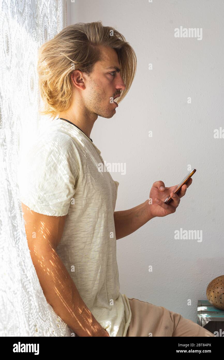 Young man with medium long blonde hair talking on the phone. Sun rays coming into the house through the window. Social distancing and technologies Stock Photo