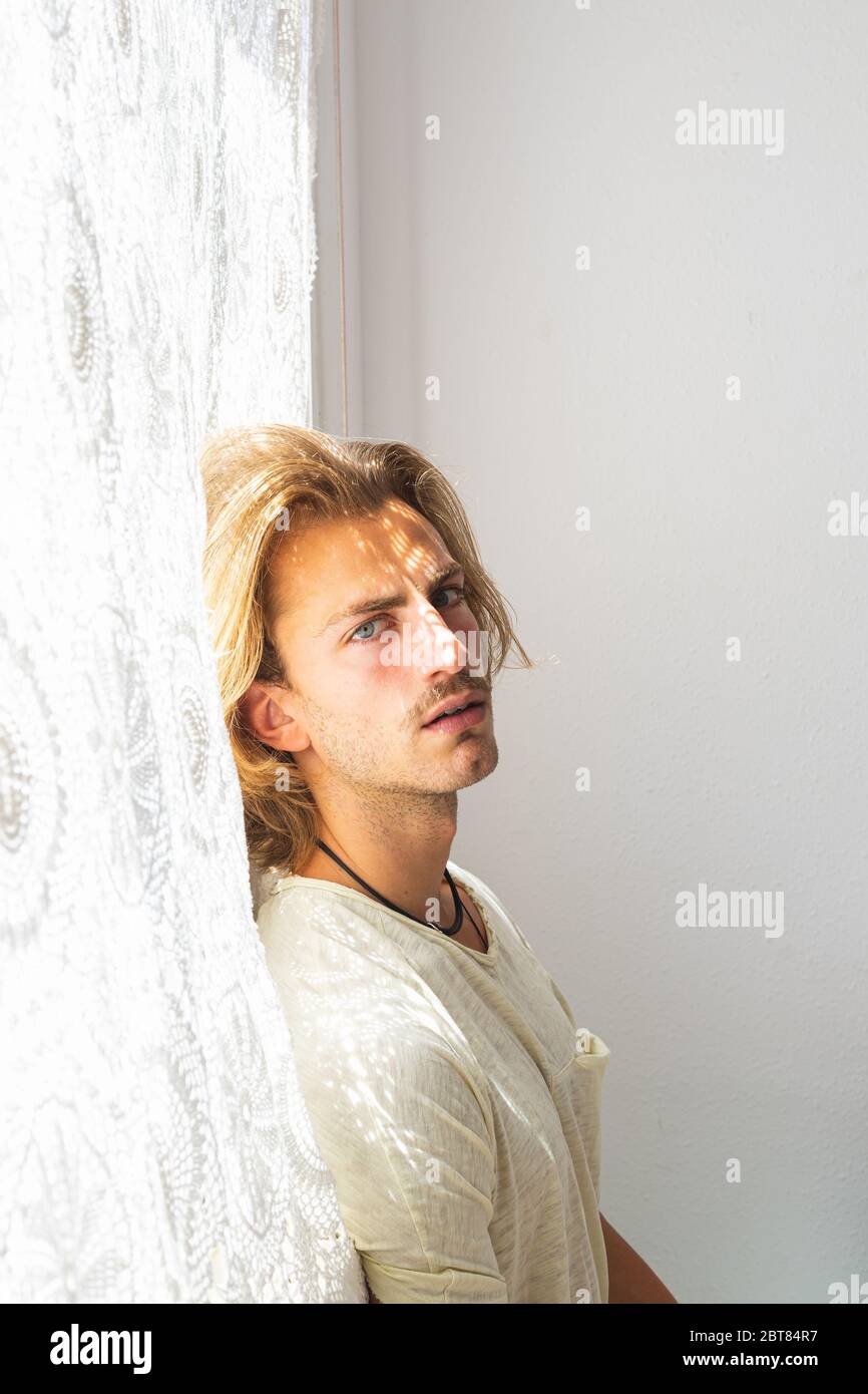 Young man with medium long blonde hair frustrated at not being able to leave home. Loneliness and silence concept. Sun rays coming into the house thro Stock Photo