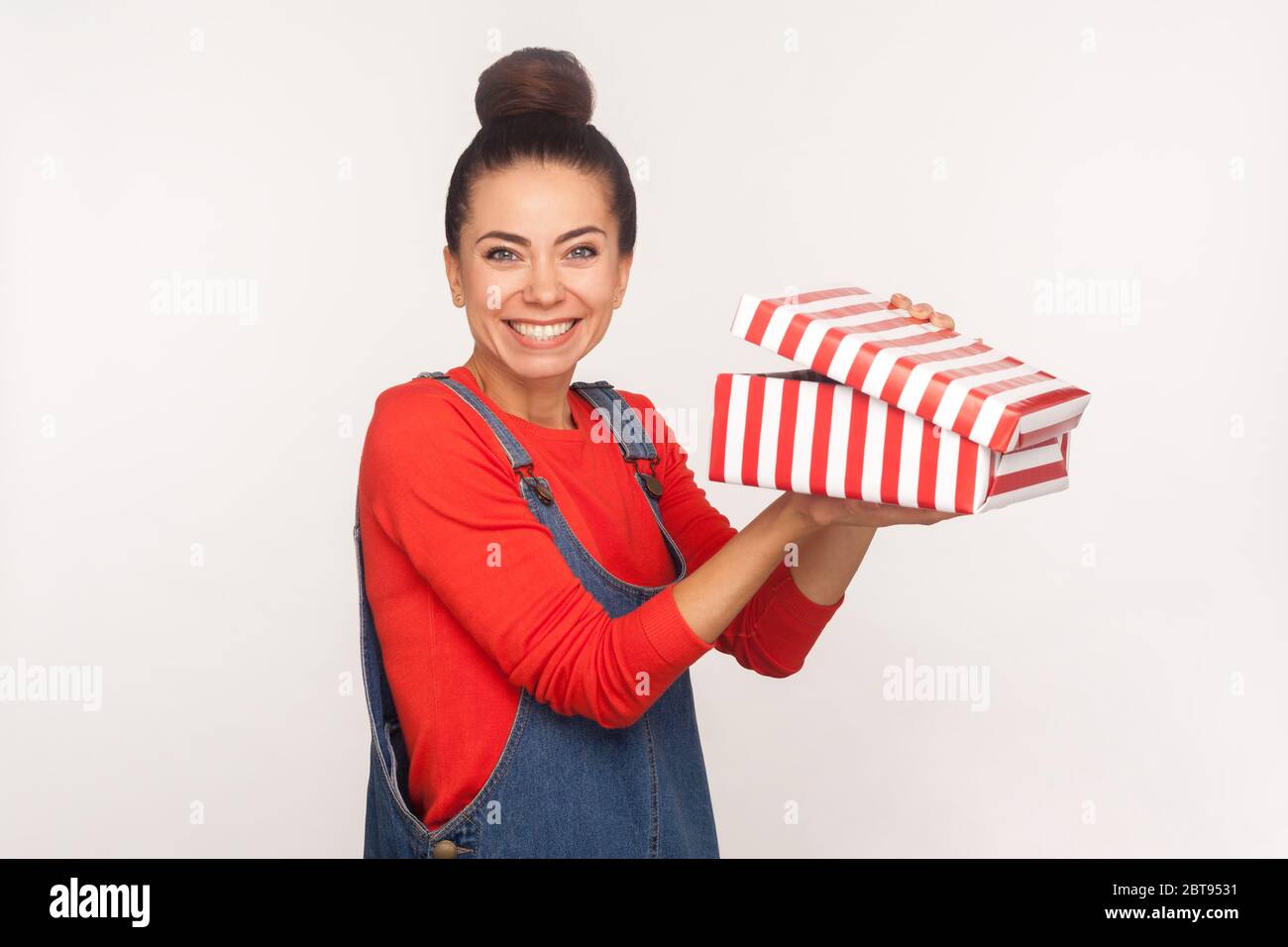 Portrait of happy pretty girl in denim overalls holding unwrapped box and smiling at camera, enjoying holiday gift, satisfied with birthday present. i Stock Photo