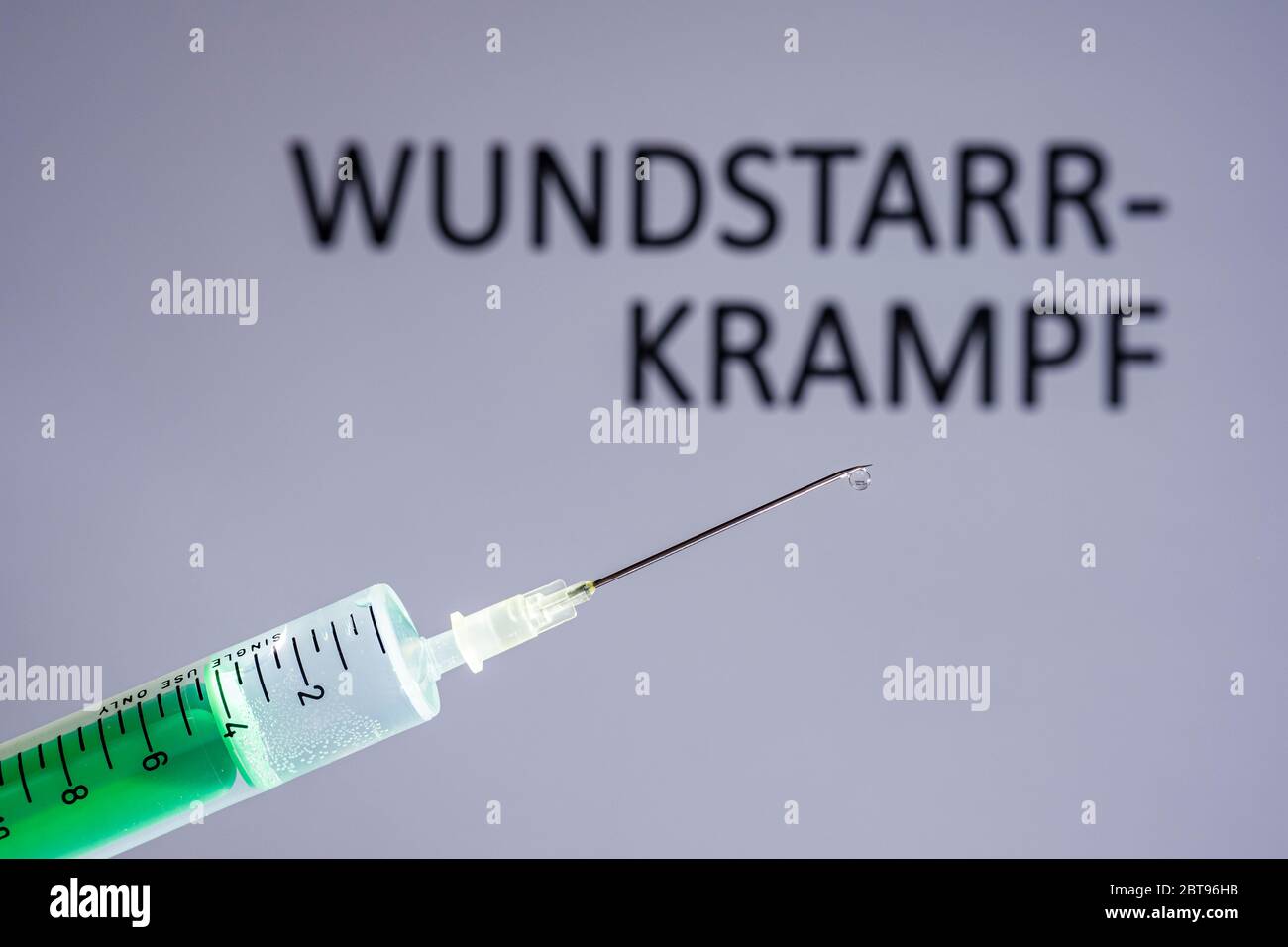 This photo illustration shows a disposable syringe with hypodermic needle, WUNDSTARRKRAMPF written on a grey board behind Stock Photo