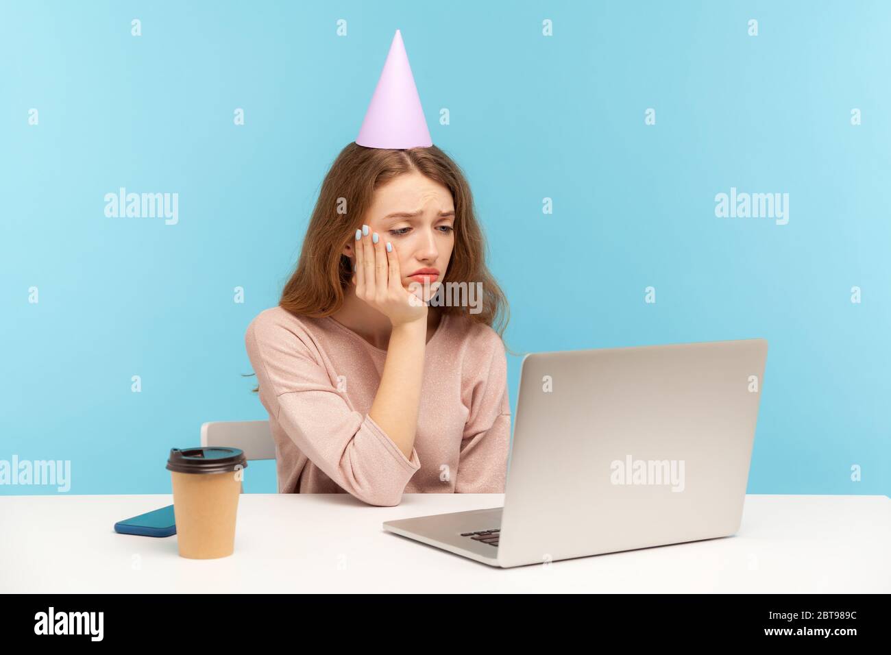 Upset woman with party cone looking sadly at laptop screen on video call and expressing boredom, frustration about lonely quarantine birthday party. i Stock Photo