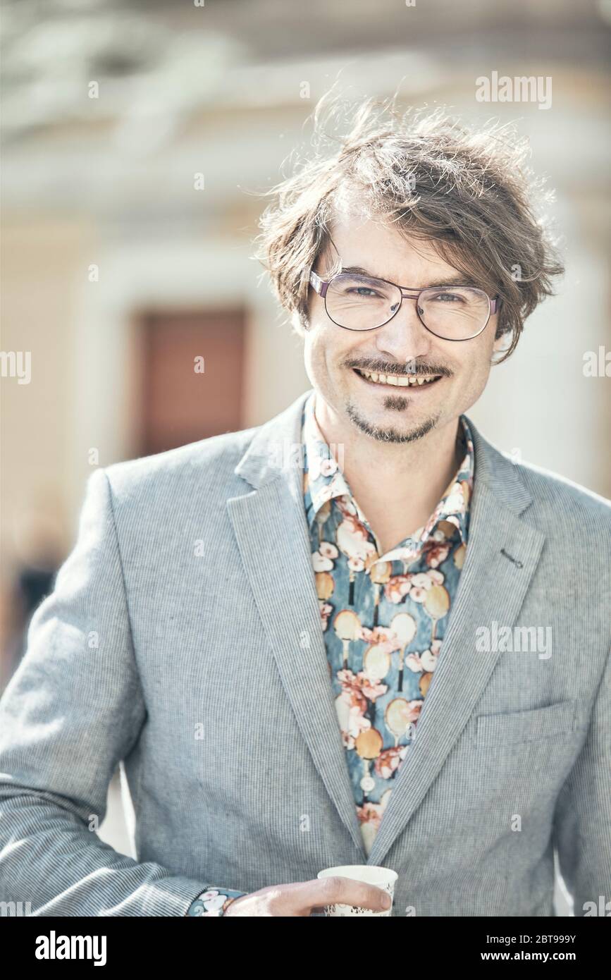 A young fashionable man closeup portrait while he is walking a street, backlit by sun, smiling to camera with spectacles, flower shirt blazer Stock Photo