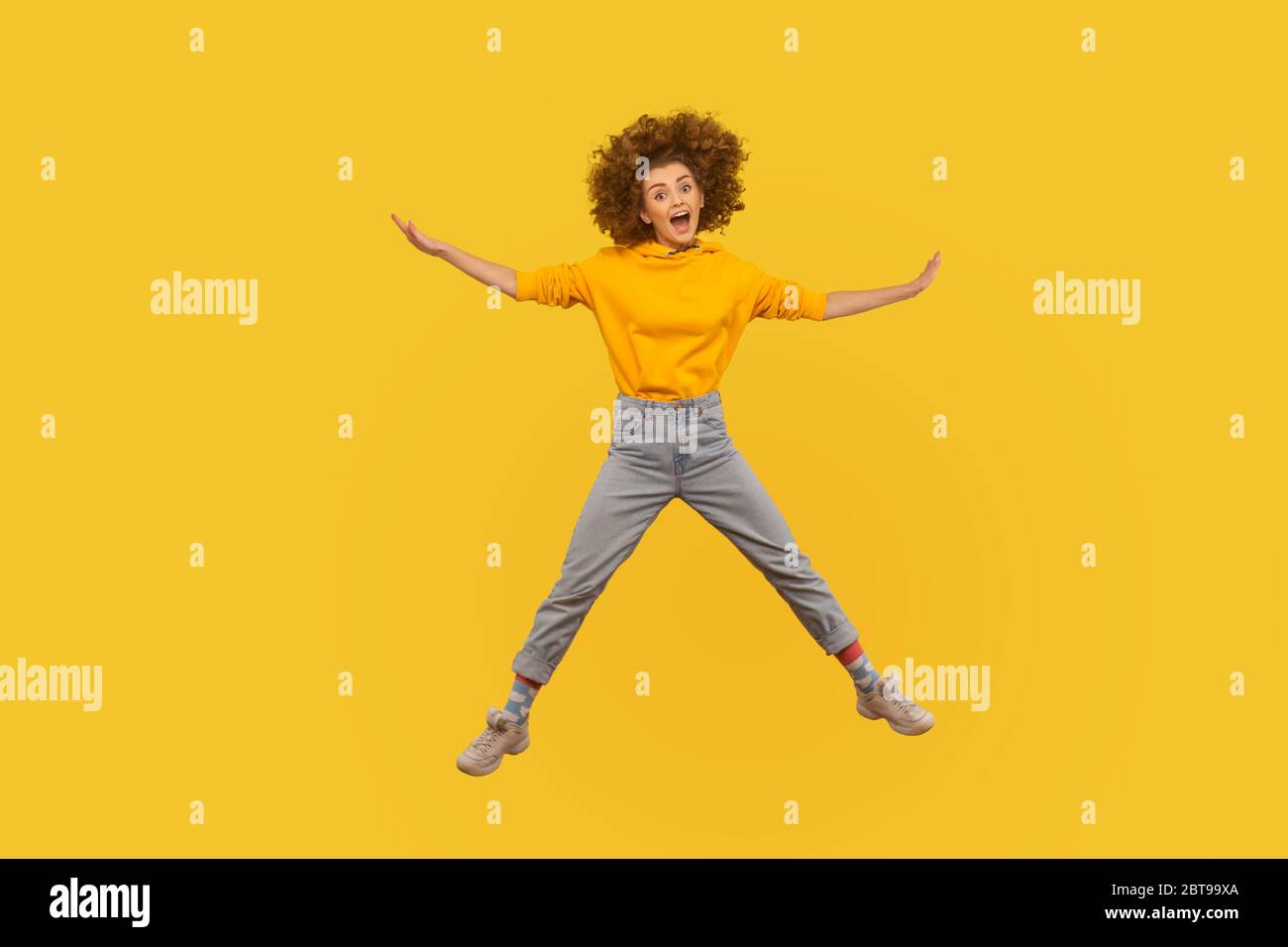Portrait of enthusiastic excited curly-haired girl in urban style hoodie and jeans jumping high in air, flying and shouting with amazed happy expressi Stock Photo