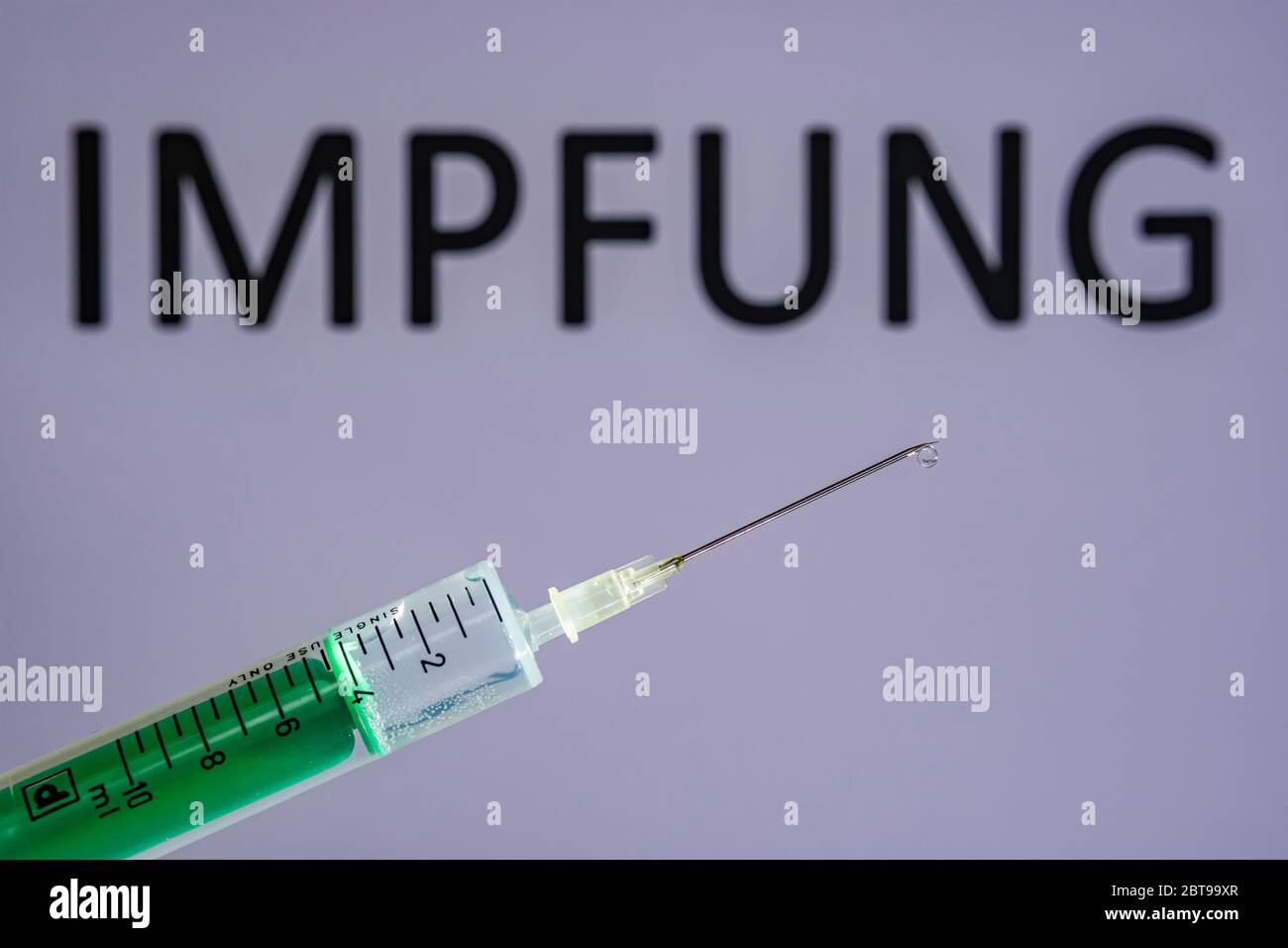 This photo illustration shows a disposable syringe with hypodermic needle, IMPFUNG written on a grey board behind Stock Photo