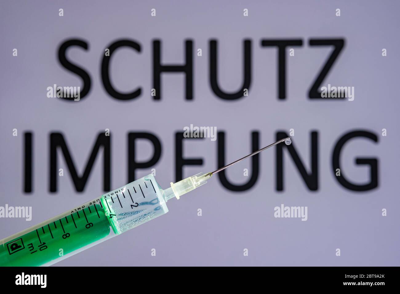 This photo illustration shows a disposable syringe with hypodermic needle, SCHUTZIMPFUNG written on a grey board behind Stock Photo