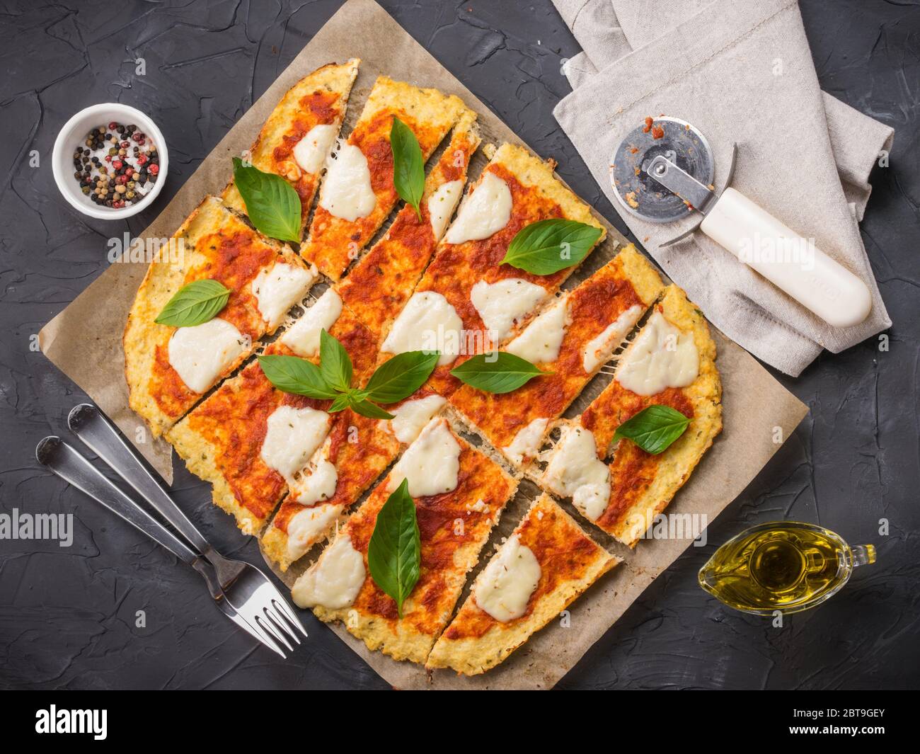 Top view of sliced homemade cauliflower pizza crust with mozarella and fresh basil in dripping pan on black concrete background. Stock Photo