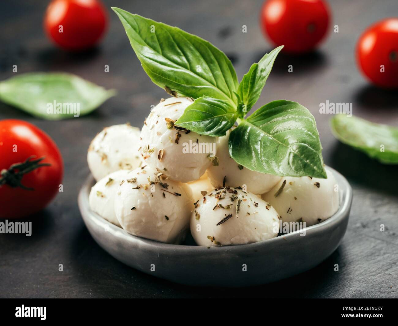 Close up view of small mozzarella cheese marinated with herbs. Mozarella balls with fresh basil and cherry tomatoes on black concrete background. Low key. Stock Photo