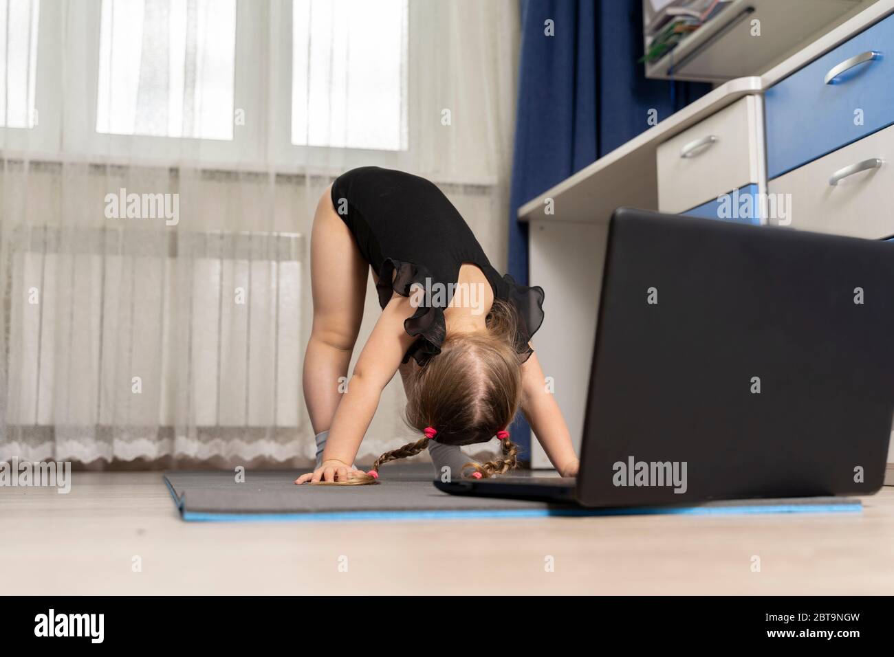 a little girl in a black gymnastics leotard is doing gymnastics at home online in front of a laptop. distance rhythmic gymnastics for children Stock Photo