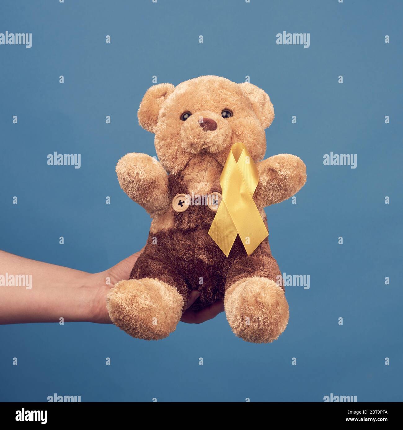 female hand hold a small teddy bear with a yellow ribbon folded in a loop on a blue background. concept of the fight against childhood cancer. problem Stock Photo