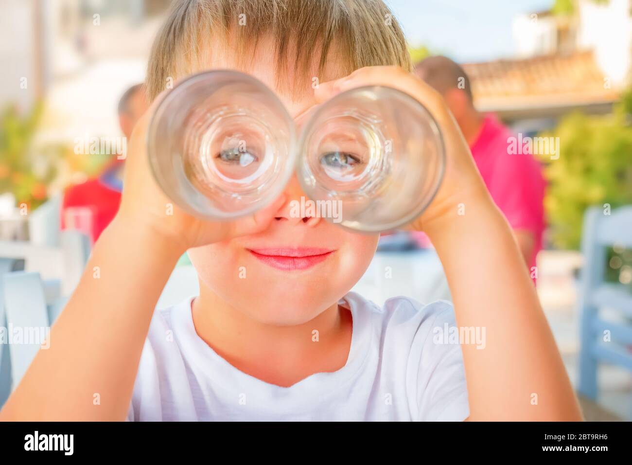 Funny kid looking through two drink glasses as if it were binoculars. Child having fun while waiting for order at cafe, restaurant. Happiness and Stock Photo