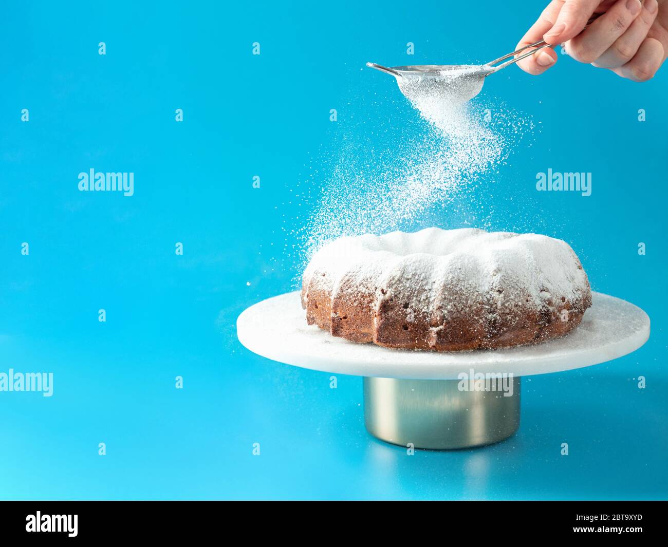 Woman's hand sprinkling icing sugar over fresh home made bundt cake. Powder sugar falls on fresh perfect bunt cake over blue background. Copy space for text. Ideas and recipes for breakfast or dessert Stock Photo