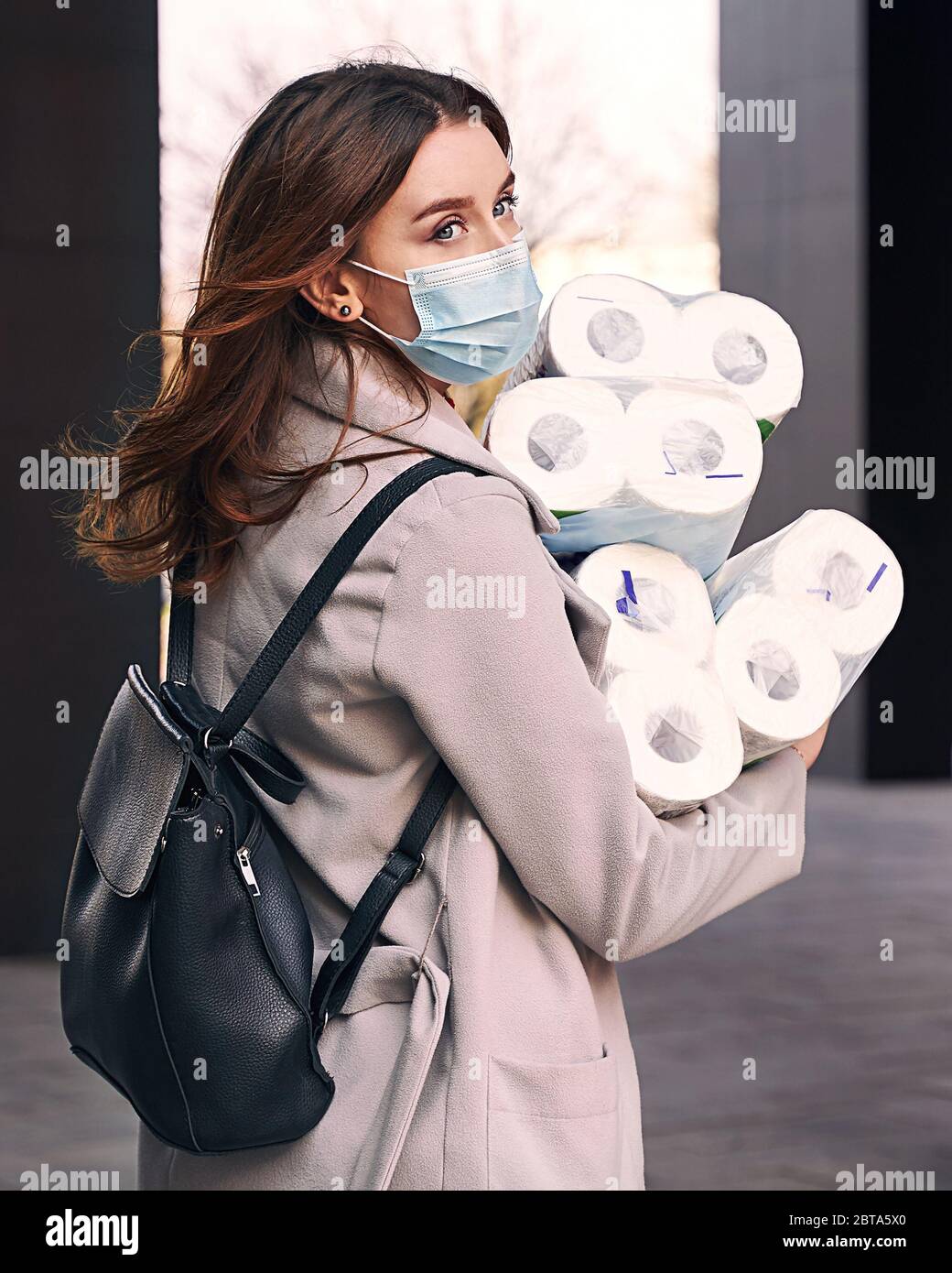Young woman wearing protection face mask and sunglasses against coronavirus COVID-19 holds many bundles of toilet paper rolls on the city street Stock Photo