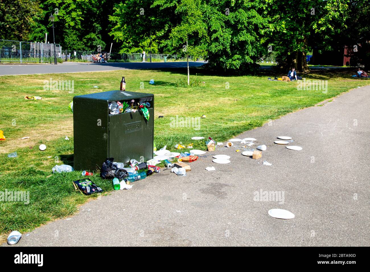 A litter bin in a park overflowing and full of rubbish, rubbish scattered on the lawn and path (Battersea Park, London, UK) Stock Photo