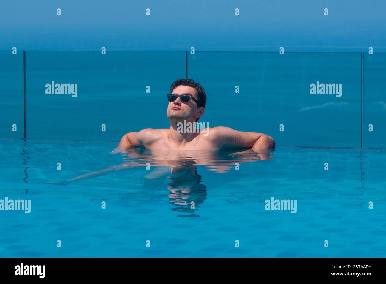 Young caucasian man wearing sunglasses at the edge of an infinity pool with blue sea and sky in the background. Royalty free stock photo. Stock Photo