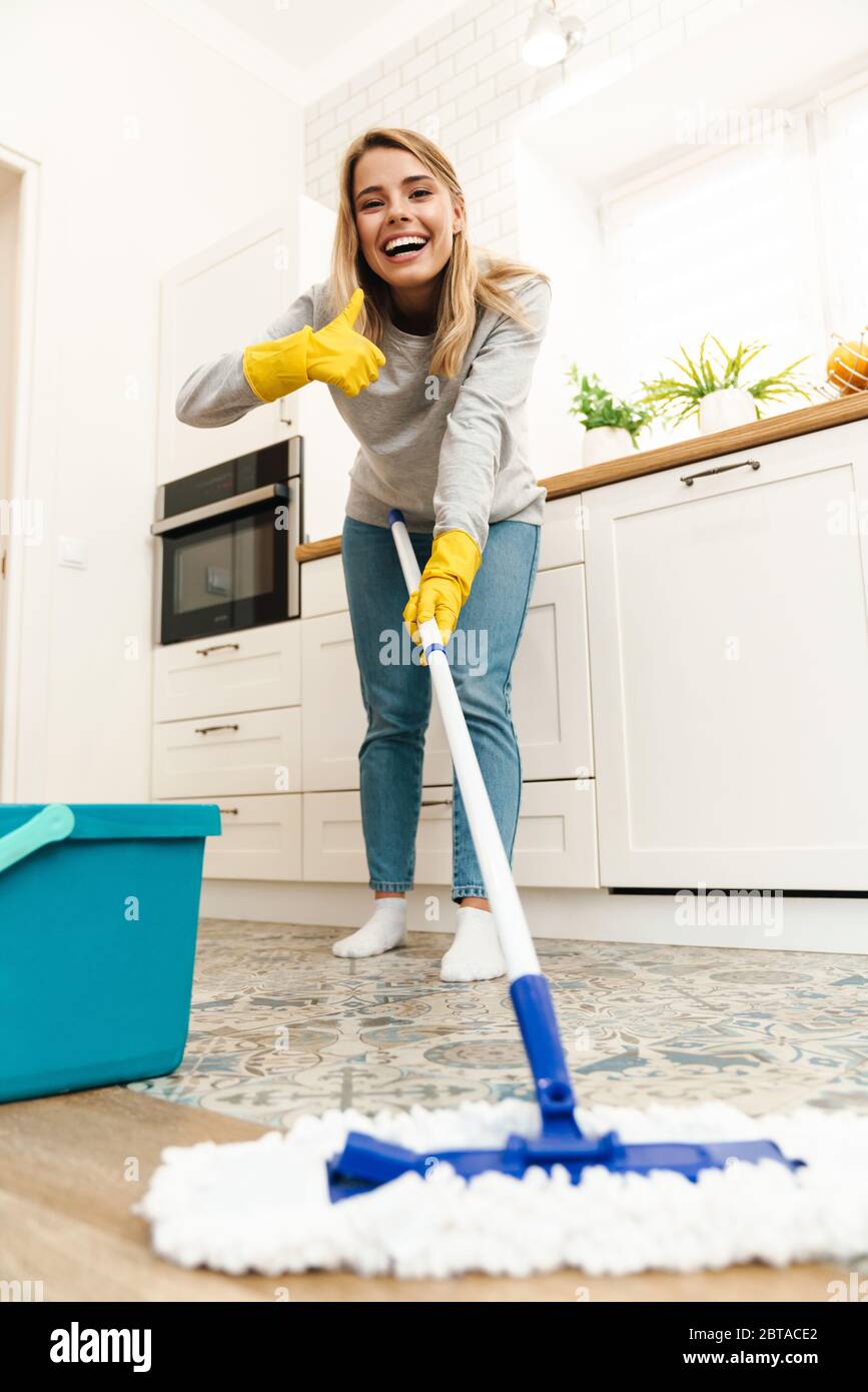 Photo of joyful young woman housewife in gloves gesturing thumb up and winking while mopping floor at kitchen Stock Photo