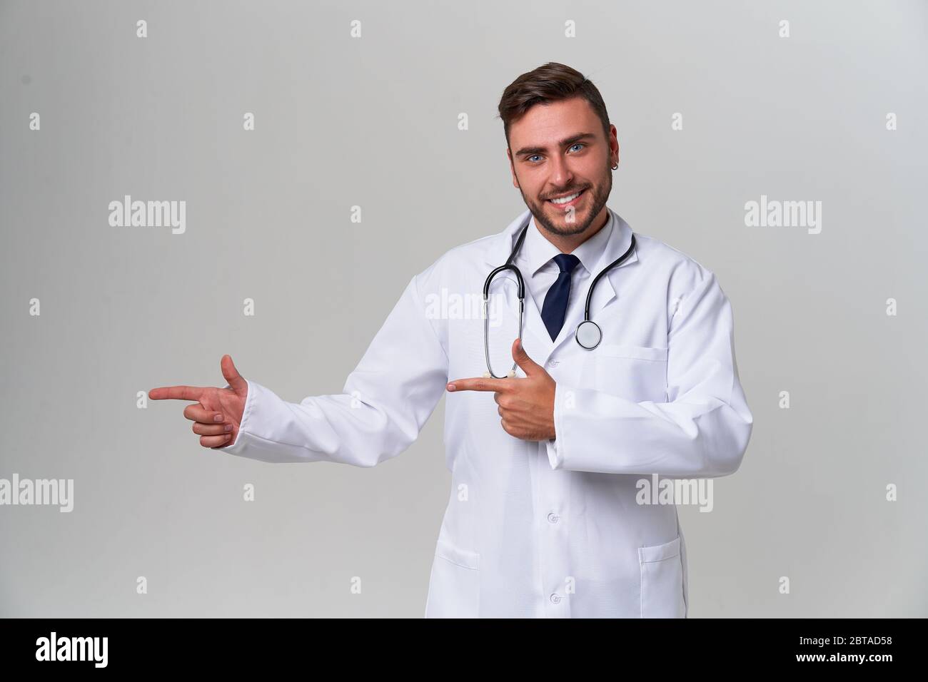 Handsome modern doctor white medical gown stands in studio gray background advertising your product looking at camera and pointing finger away Positiv Stock Photo
