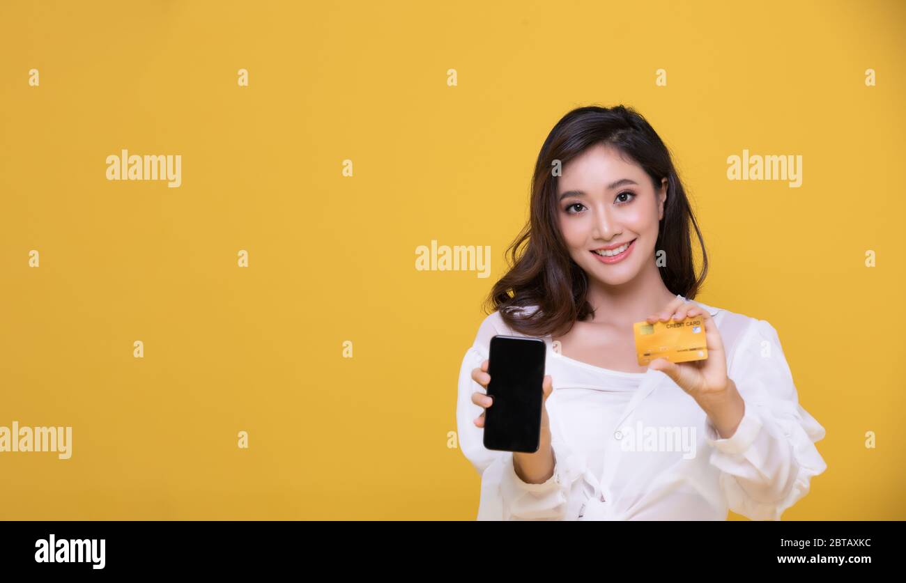 Portrait Asian beautiful happy young woman smiling cheerful and she is holding credit card and using smart phone for shopping online on yellow backgro Stock Photo