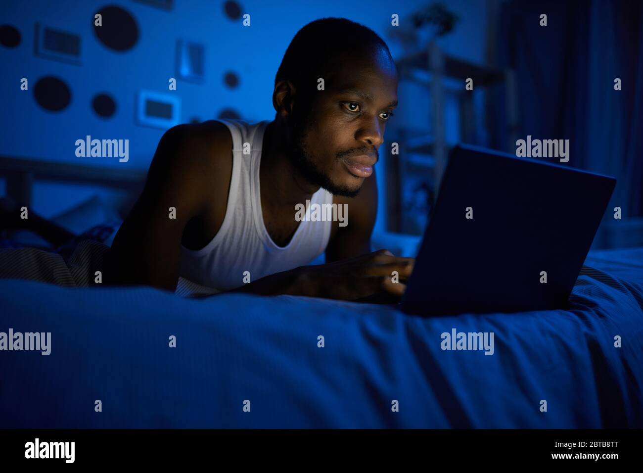 Portrait of young African-American man using laptop while lying on bed at night, copy space Stock Photo